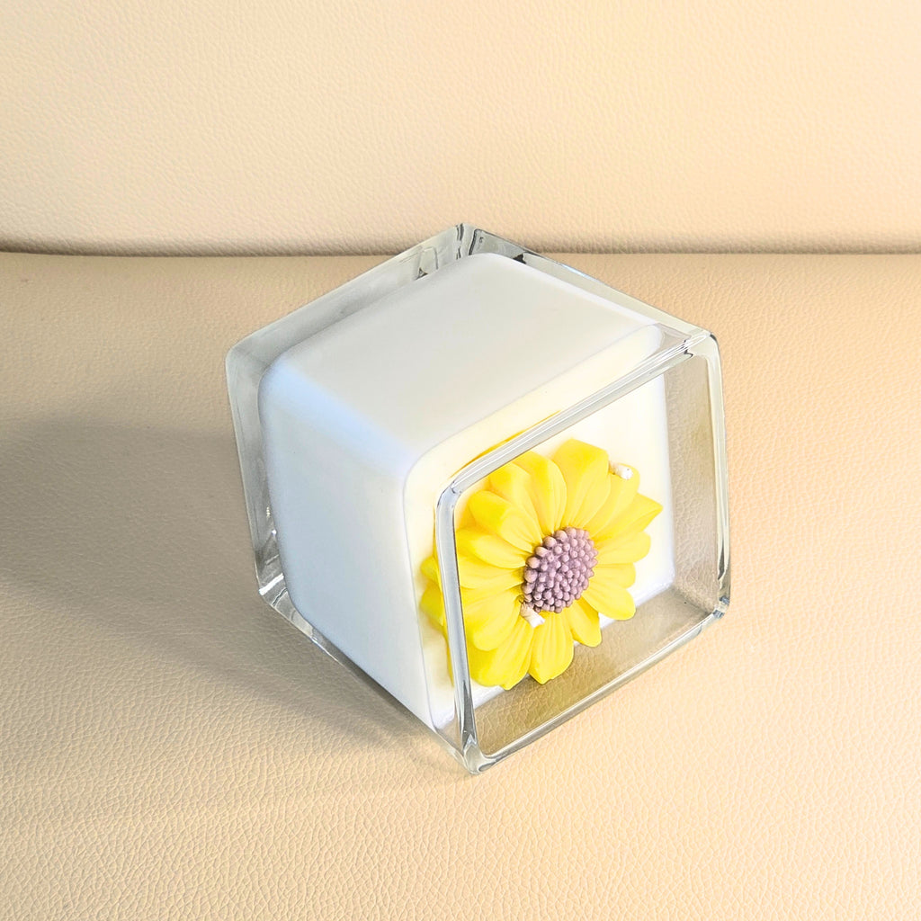 Side angle of luxury yellow-flower candle in a clear square glass vessel.