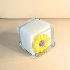 Side angle of luxury yellow-flower candle in a clear square glass vessel.